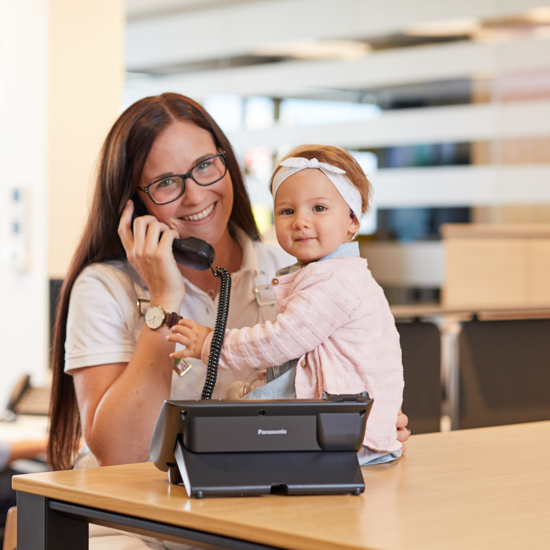 Mitarbeiterin telefoniert mit Kinderwunsch-Patientin, Kleinkind sitzt daneben – persönlicher Kontakt zur Kinderwunsch-Beratung
