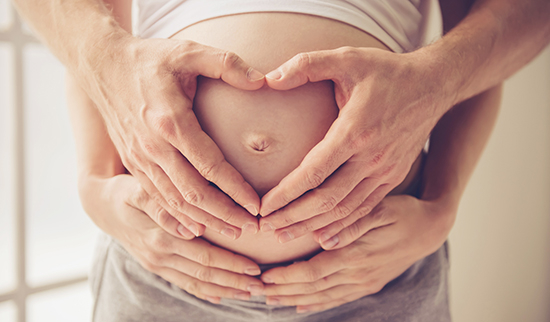 Schwangere Frau mit eigenen Händen und den Händen ihres Partners am Bauch – Symbolbild für Schwangerschaft und Kinderwunsch